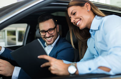 Photo of female smiling and presenting sales number to a male in the drivers seat of a vehicle